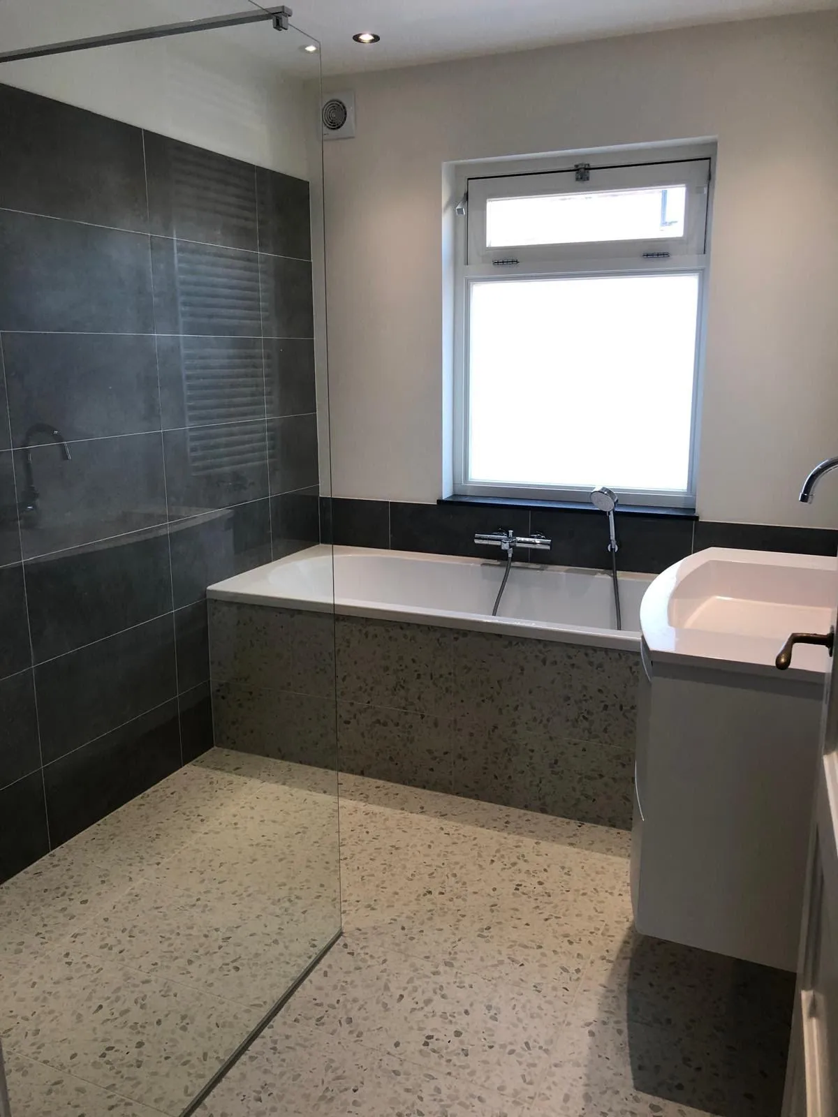 Badkamer met inbouwbad, donkere wandtegels, terrazzovloer, glazen douchewand en raam
