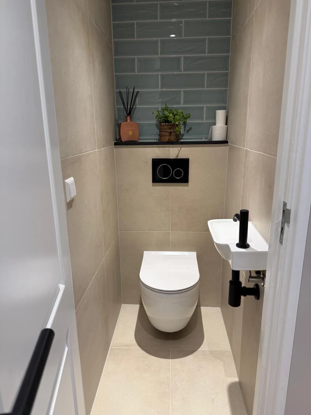Gerenoveerd toilet met hangend closet, zwarte bedieningsplaat, groen betegelde nis en klein fonteintje